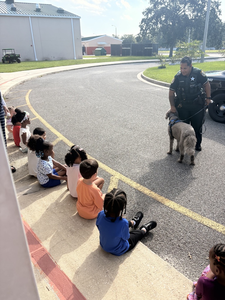 Children seated on a curb watch as a police officer walks a dog nearby on a street.