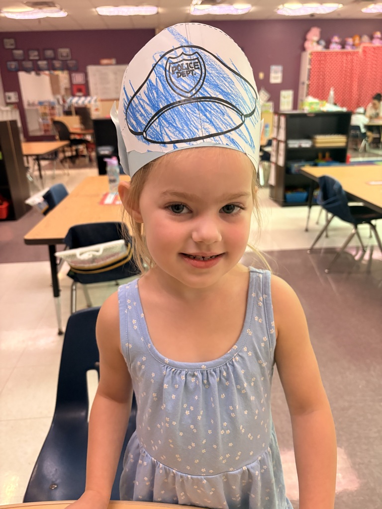 A child stands in a classroom wearing a paper hat shaped like a police officer's hat.