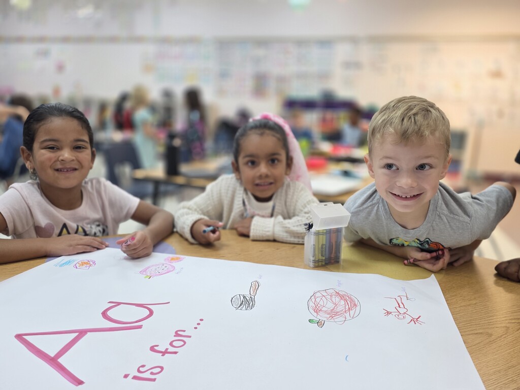 Three children sit at a table, smiling with a paper saying "Aa is for...".