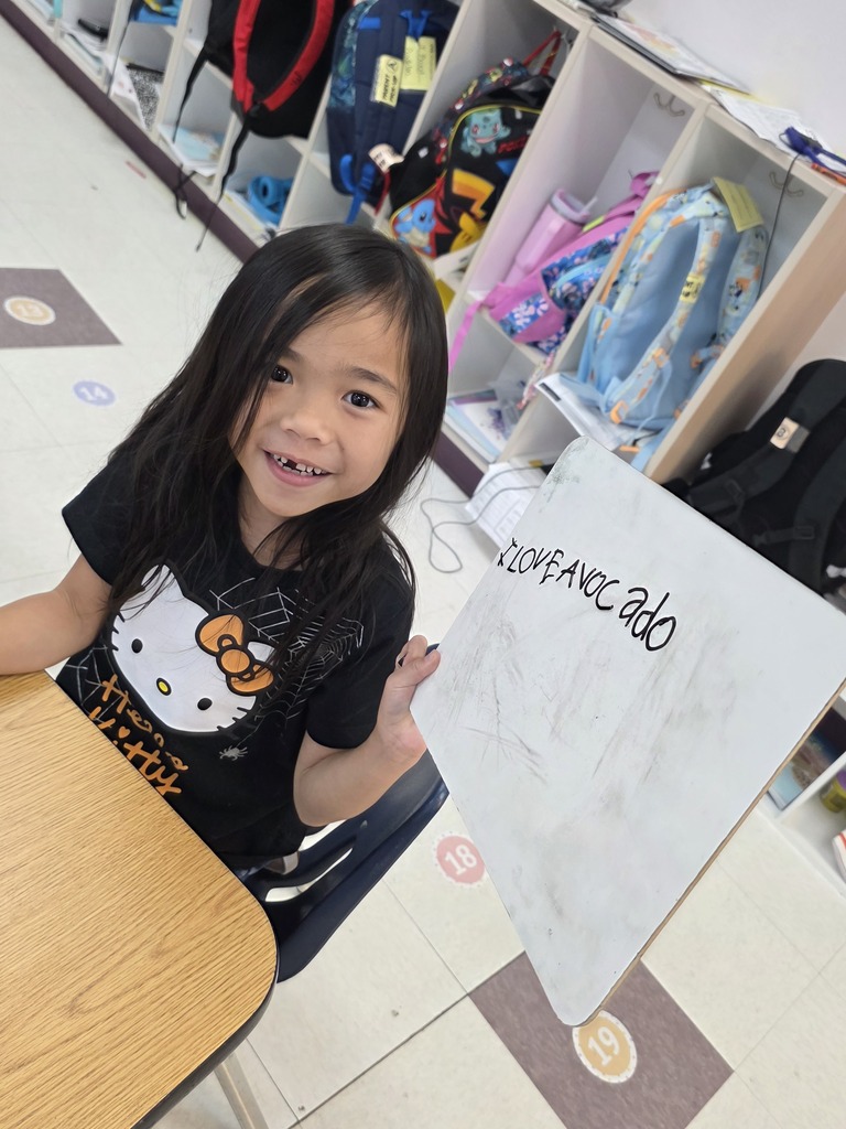 A young girl smiles and holds a whiteboard with writing while sitting at a desk. 