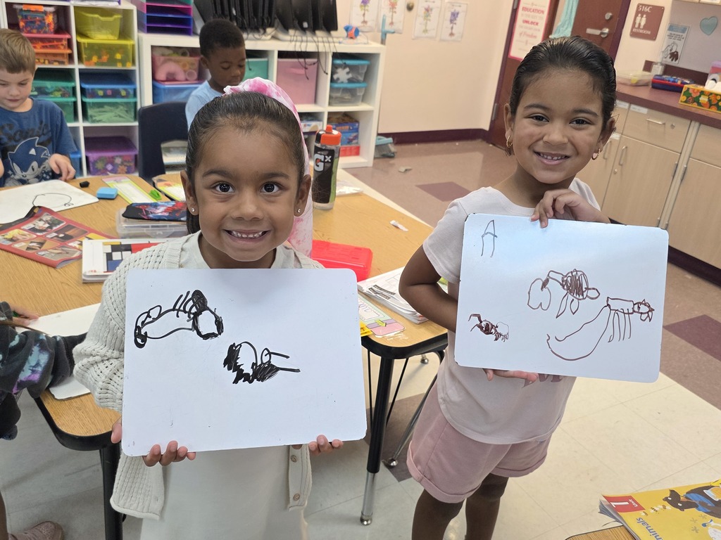 Two young girls in a classroom hold up drawings, smiling. Behind them, more children are at desks.