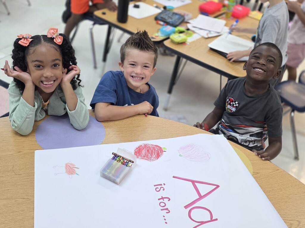 Three children sit at a table with a poster reading "A is for..." 