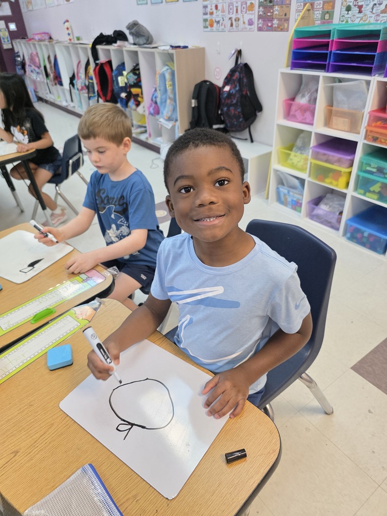 Two children in a classroom, one sitting, smiling, and drawing on a whiteboard, the other seated with a marker.