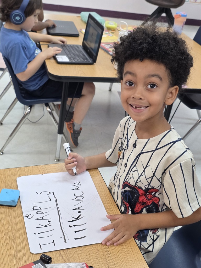 A young boy, smiling, holds a marker and whiteboard with words at a table.