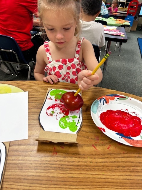 A young girl paints an apple on a cardboard cutout with a brush. She sits at a table with other art materials.