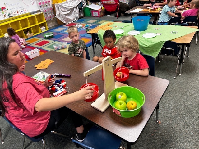 Woman sits at a table with two children. They play with a toy fruit stand. Other children sit at tables in the background.