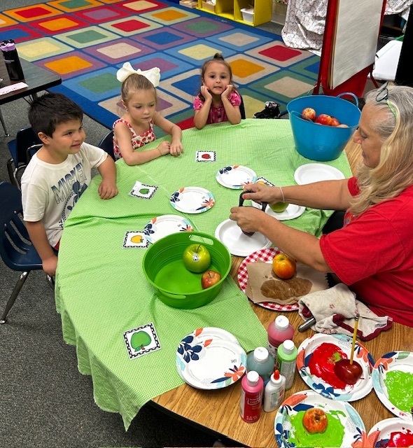 Children and an adult at a table, with an adult preparing apples. Bright multicolored rug.