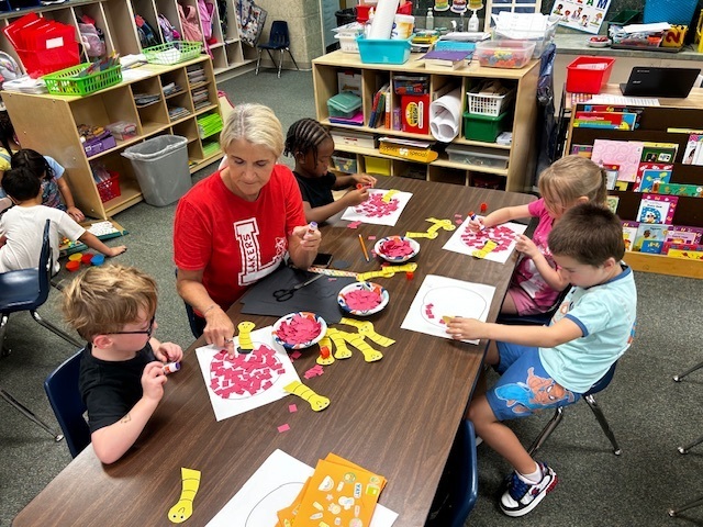 A woman leads children in a classroom, using scissors to cut shapes. The kids sit at a table, engaged in the activity.