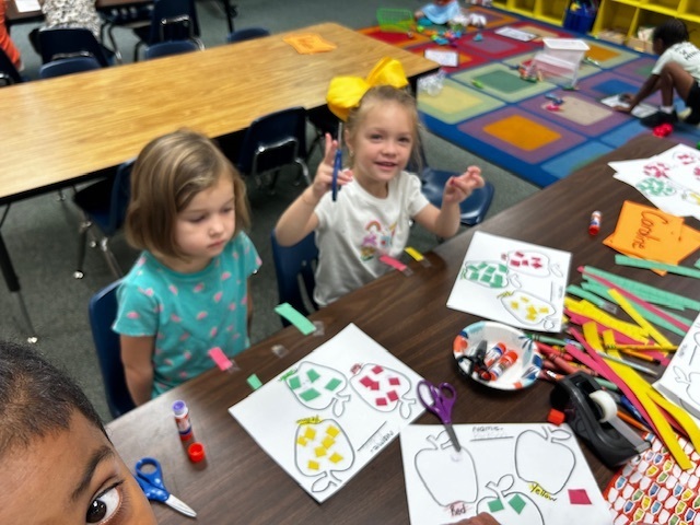 Two girls sit at a table with crafts; one holds a glue stick. One girl wears a bow, and both have papers with drawings.