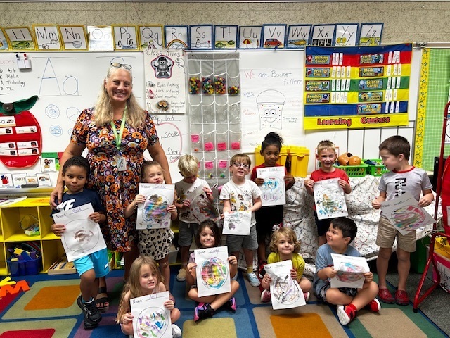 A woman stands in a classroom with children holding certificates. They are seated on the floor.