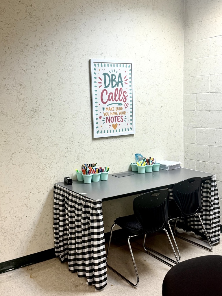 A classroom with a desk covered in a black and white checked tablecloth. Two chairs surround the desk.