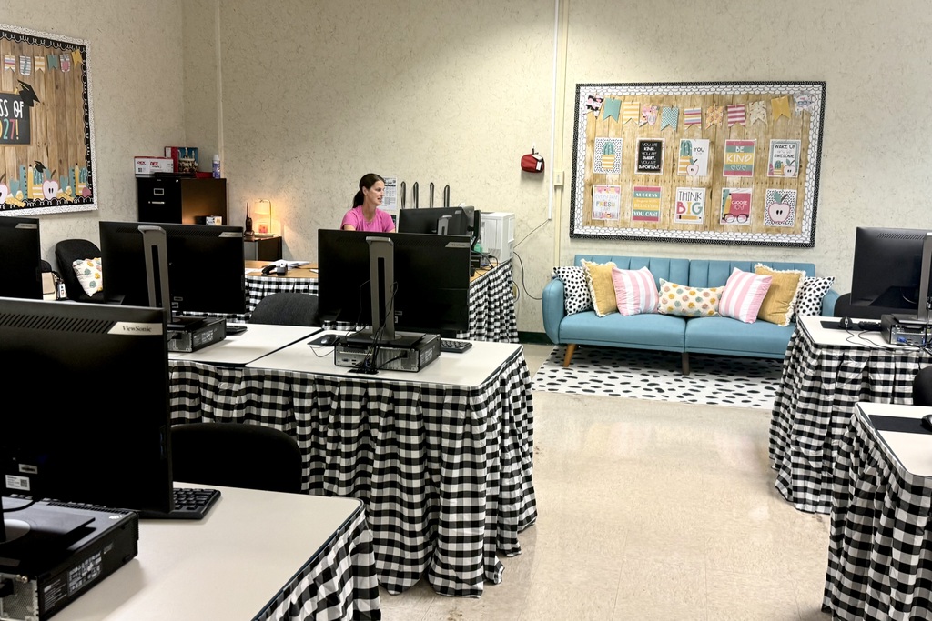 An office with computer monitors on desks, a person standing in front of a computer, a couch with pillows, and framed artwork on the wall.