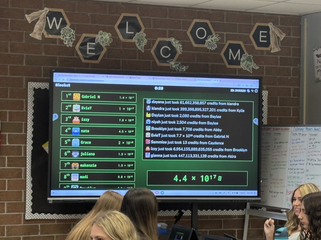 A classroom with a brick wall. A large screen displays a math problem. Students sit and listen.