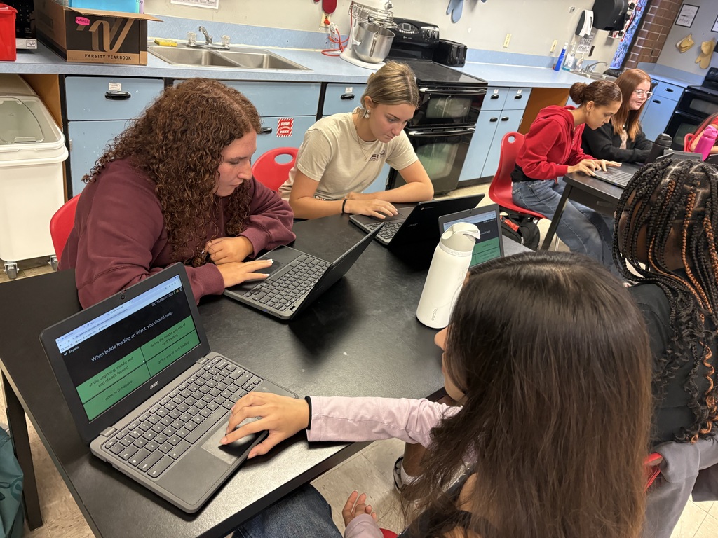 A group of students seated at a table in a classroom, each using laptops. A countertop with kitchen appliances is visible in the background.