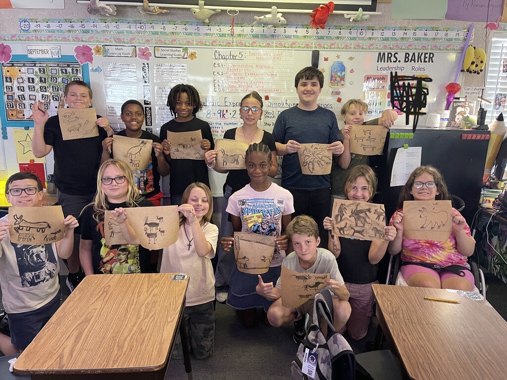 A classroom with students, including one teacher, holding drawings and posing for a group photo.