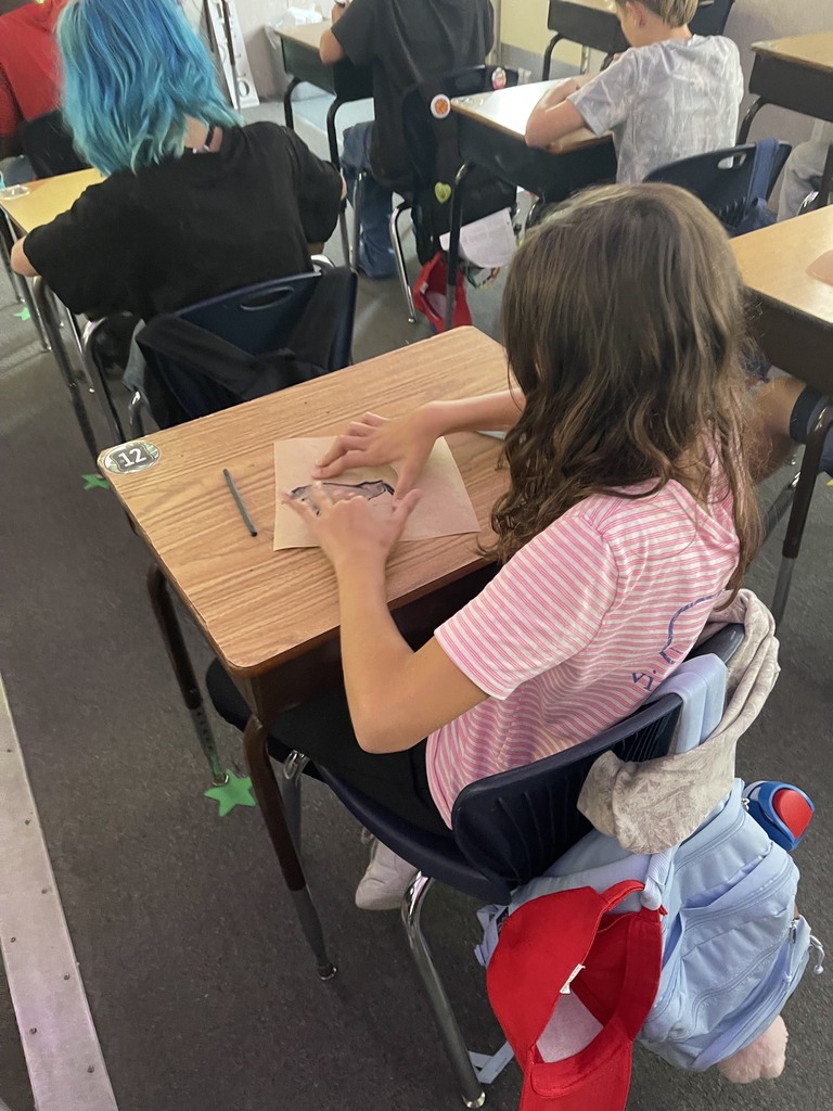 A girl with long brown hair sits in a chair in a classroom, writing on a piece of paper.