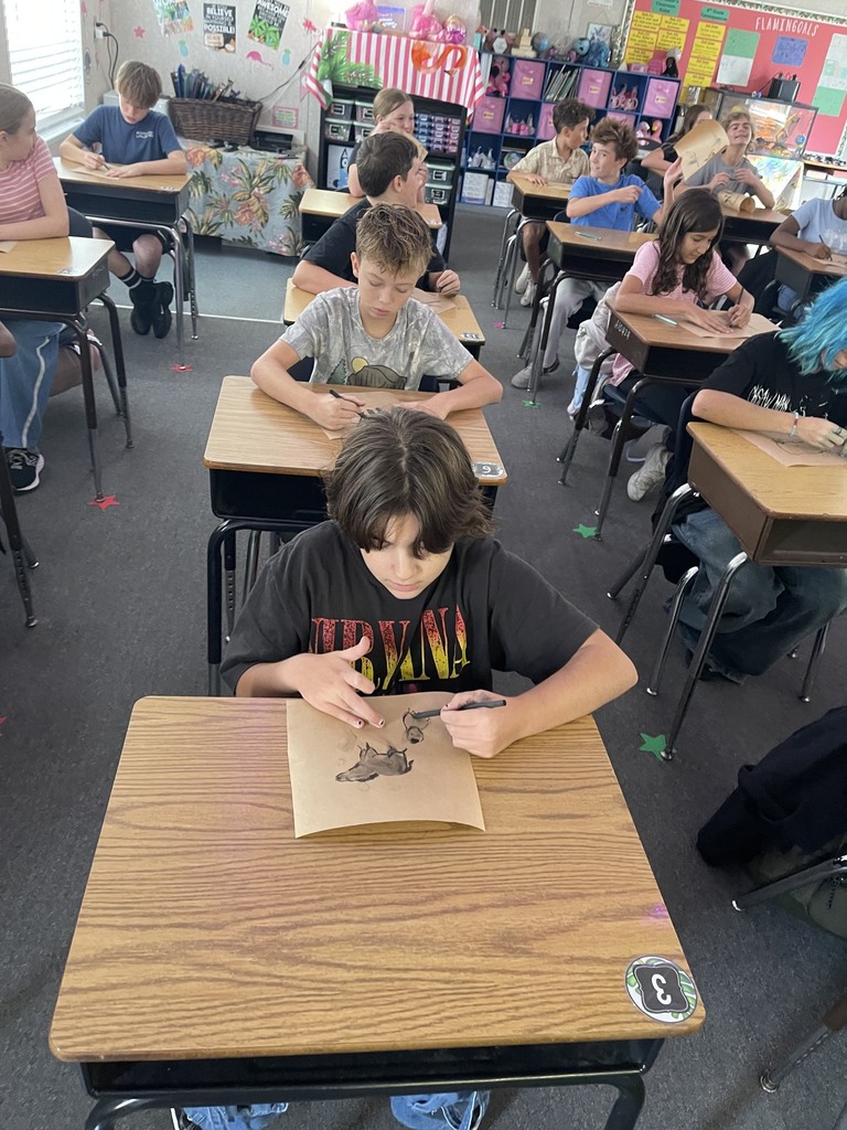 Students in a classroom are seated at desks, engaged in individual activities. One student focuses on a paper, while others write or draw.