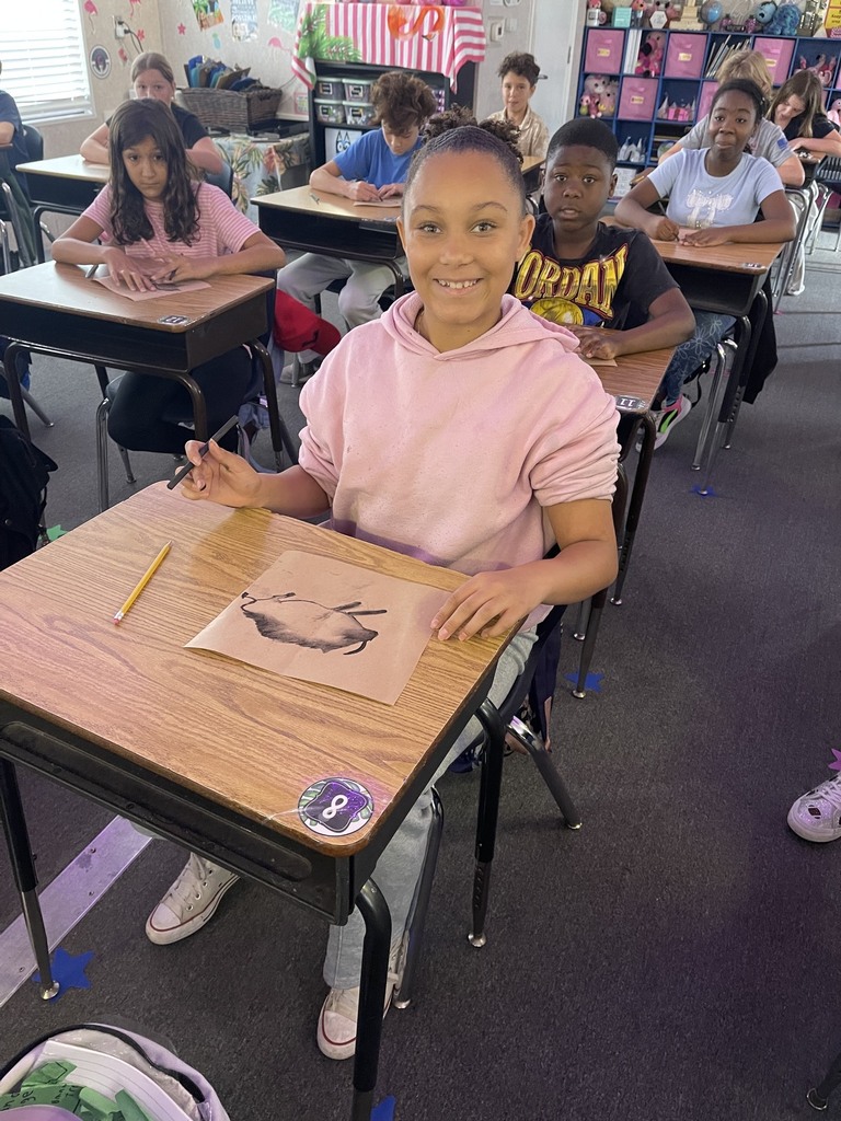 A student in a pink hoodie draws on paper at a desk in a classroom. Other students sit at desks in the background.