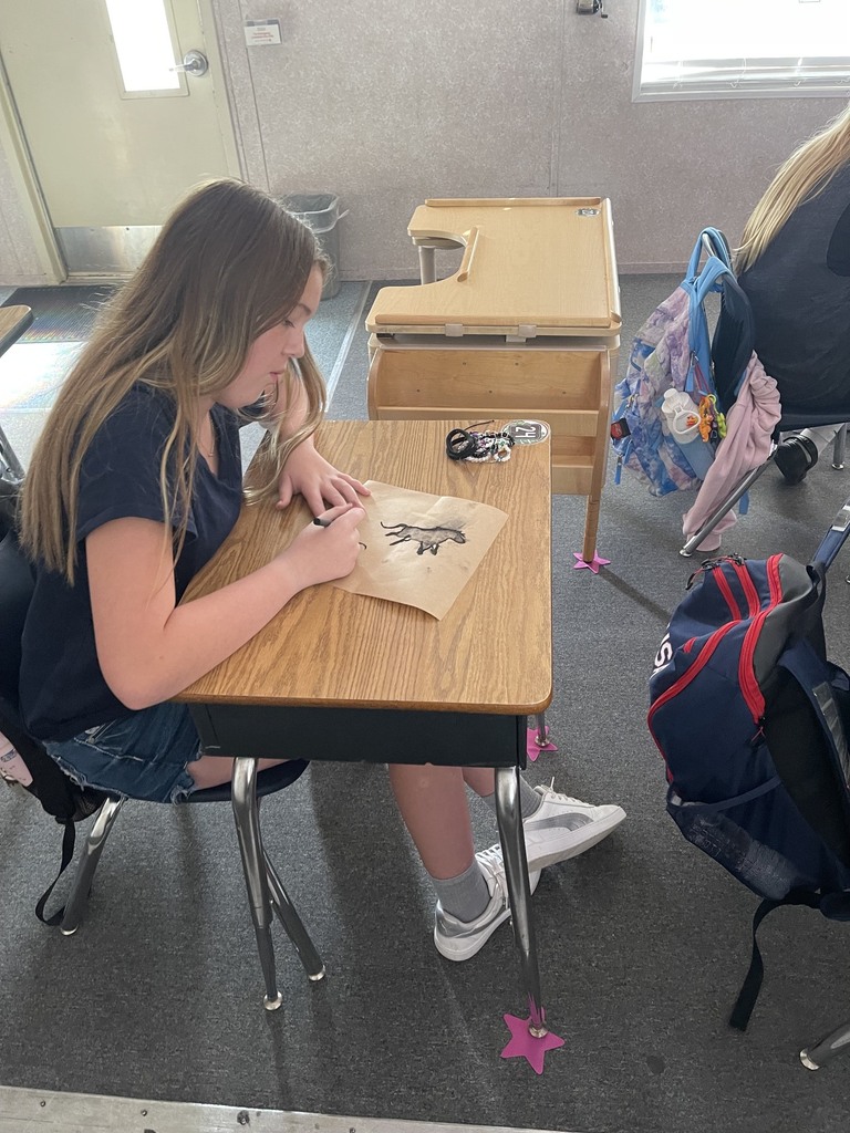 A person sits at a desk, drawing on paper. Desks and chairs are in the background.