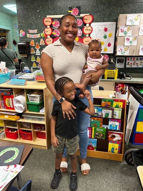 Woman holding a baby and a child in a classroom with shelves of books and other materials.