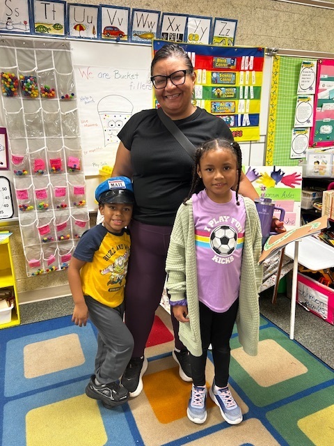 Two children stand with a woman in a classroom. They wear casual clothes. Bright colors and educational posters adorn the background.