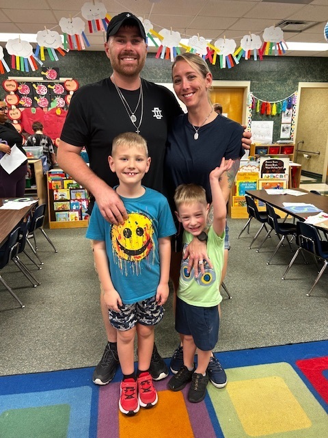 A family stands in a classroom, with two children and two adults. The room has tables, chairs, and a colorful rug.