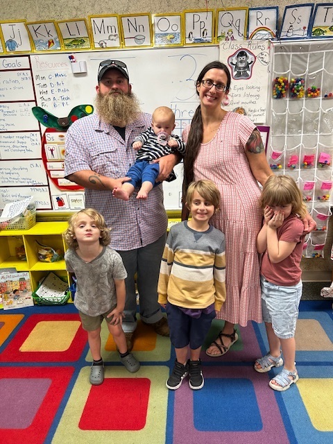 Family of five, including a baby, pose in a classroom. Two adults hold baby, while three children stand. Background includes a colorful rug, shelves, and a whiteboard.
