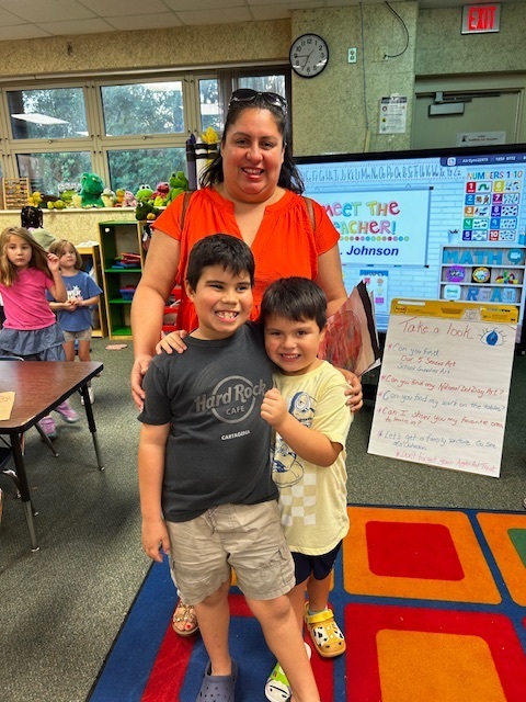 Woman stands with two boys in classroom. A colorful rug, desks, and a clock on wall in background.
