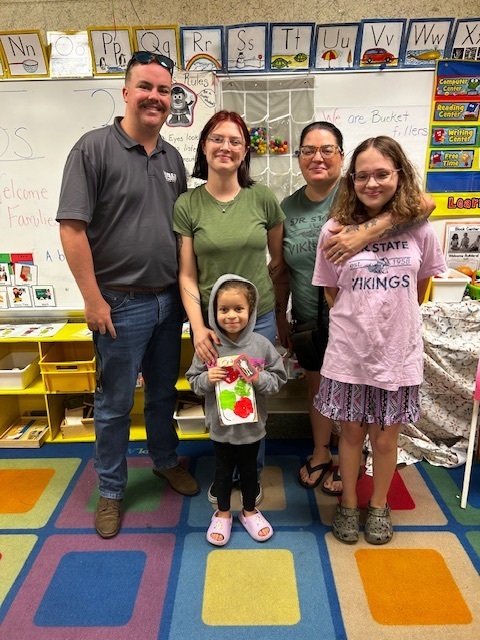 Five people stand in a classroom with multicolored floor tiles. Two women and two children wear glasses. One child holds a wrapped gift.