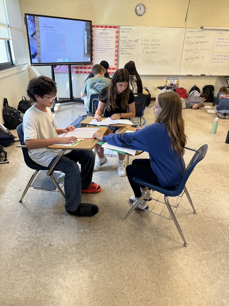 Students seated at tables, working on papers, with a monitor and whiteboards in the background.
