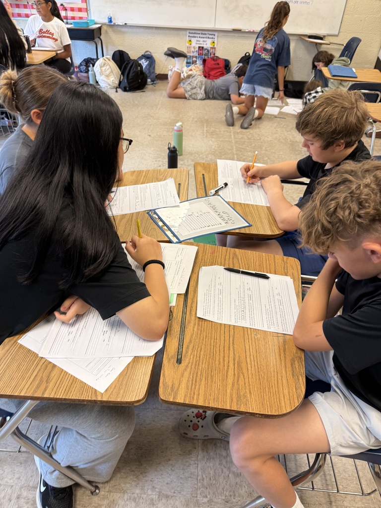 Several students are seated at desks in a classroom. One student is writing, and others have papers in front of them.