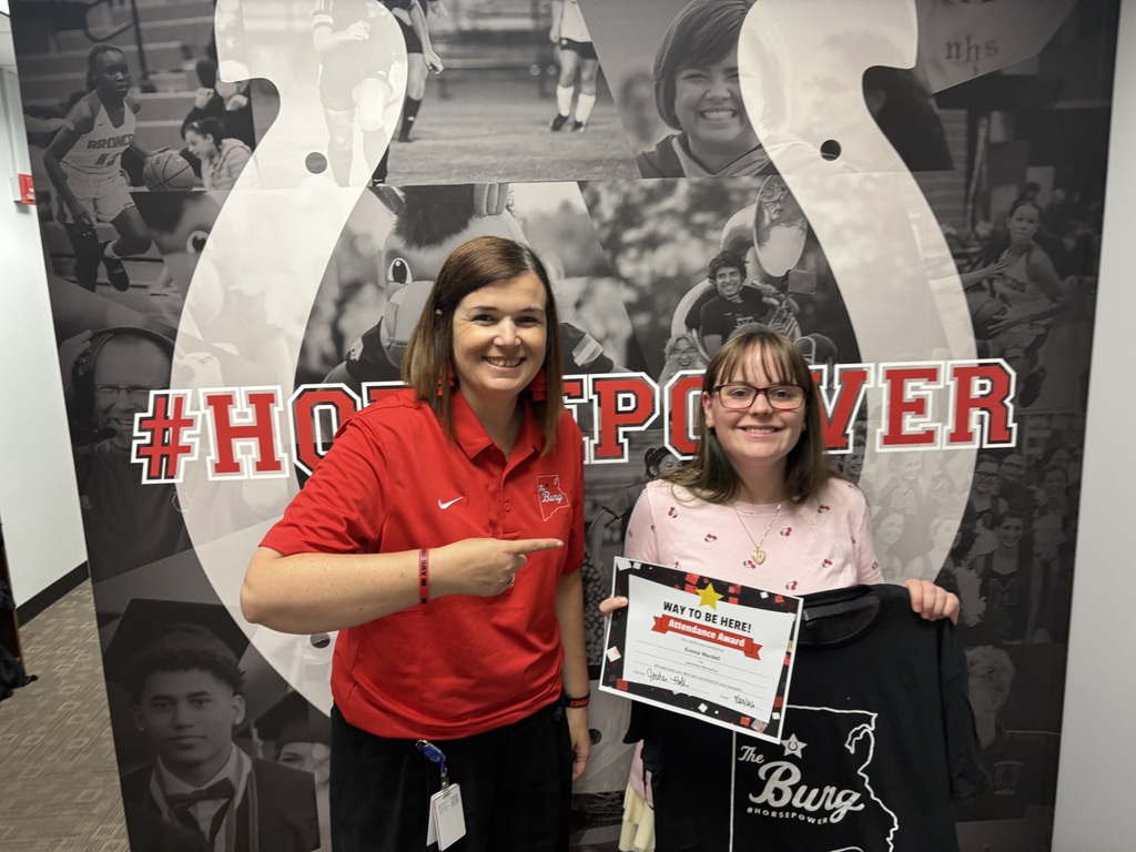 Two women stand in front of a large, decorative horseshoe with sports photos. They hold a certificate and a T-shirt.