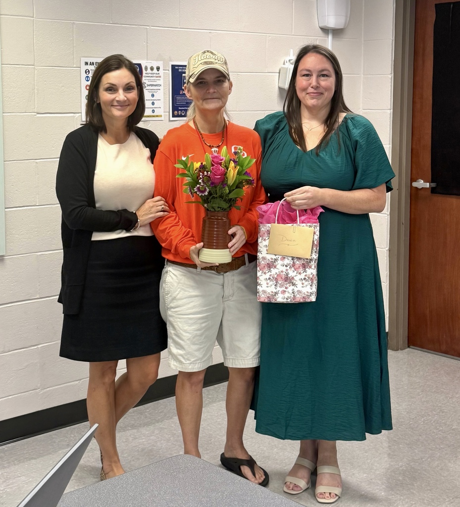 Three women standing close together in a room, one holding a flower bouquet, one in a dress holding a gift bag.