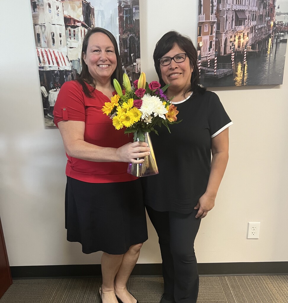 Two women standing next to each other. One holds a bouquet of flowers. Behind them are two framed paintings.