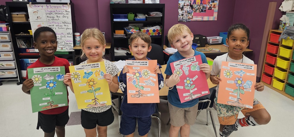 Five children stand in a classroom, holding up colorful posters with images and text. Behind them is a white board and shelves with items.
