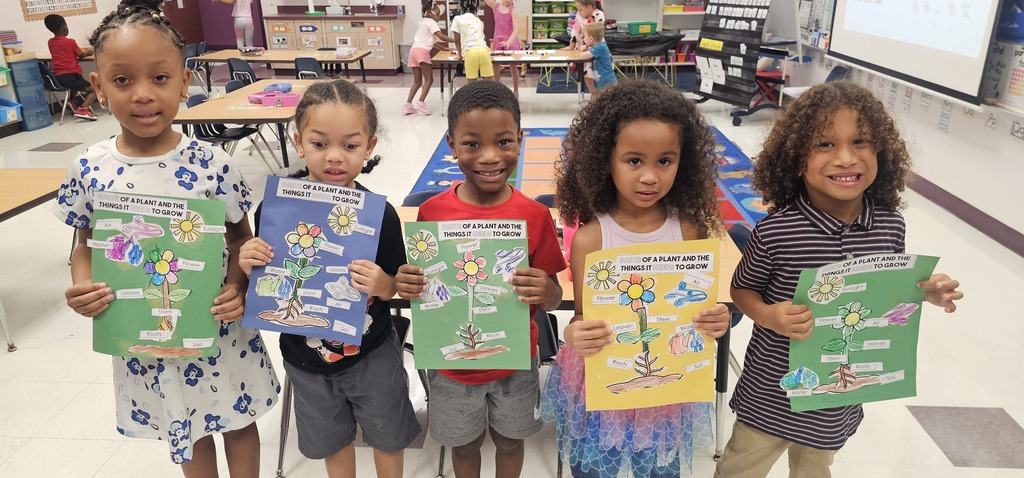 Five children in a classroom, holding up colored papers, smiling, and standing in front of tables.