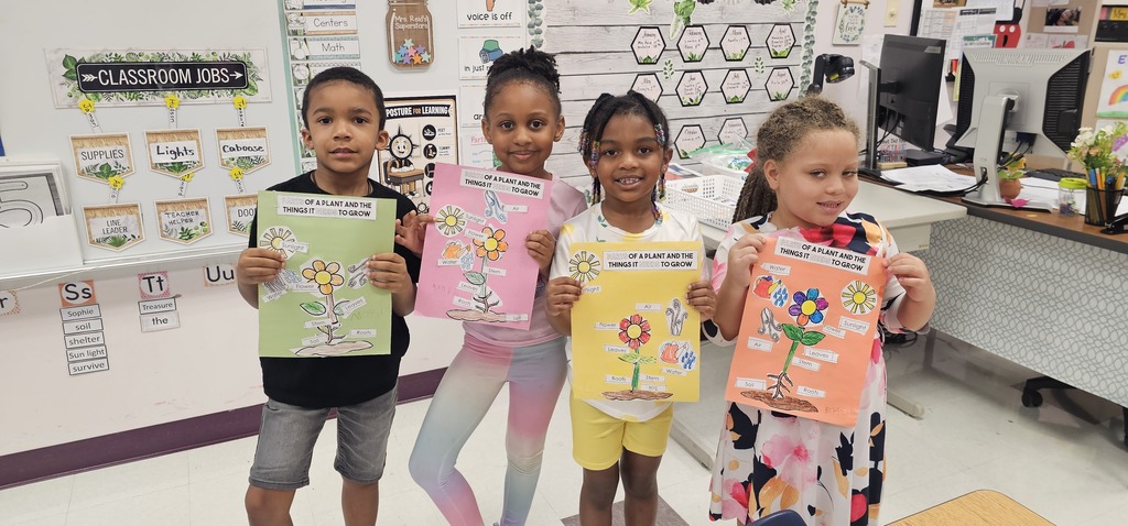Four children in a classroom, holding up colorful posters, smiling, with a white wall, posters, and a desk in the background.