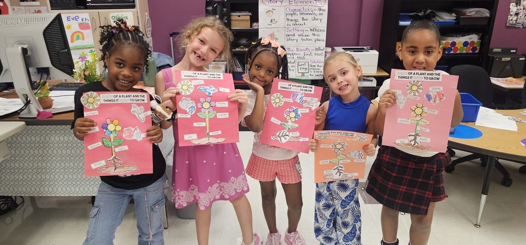 Five children in a classroom pose with pink and orange posters. A table, chair, and shelf are behind them.