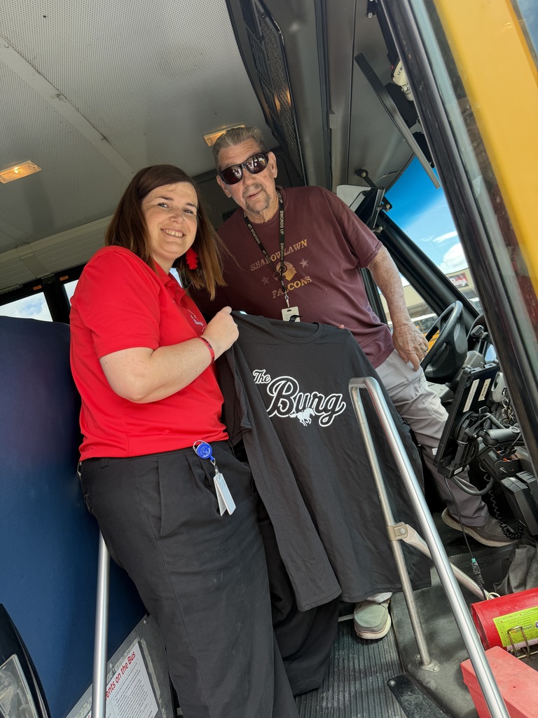 A woman in a red shirt holds a black T-shirt inside a bus. Next to her, a man in sunglasses holds the T-shirt.