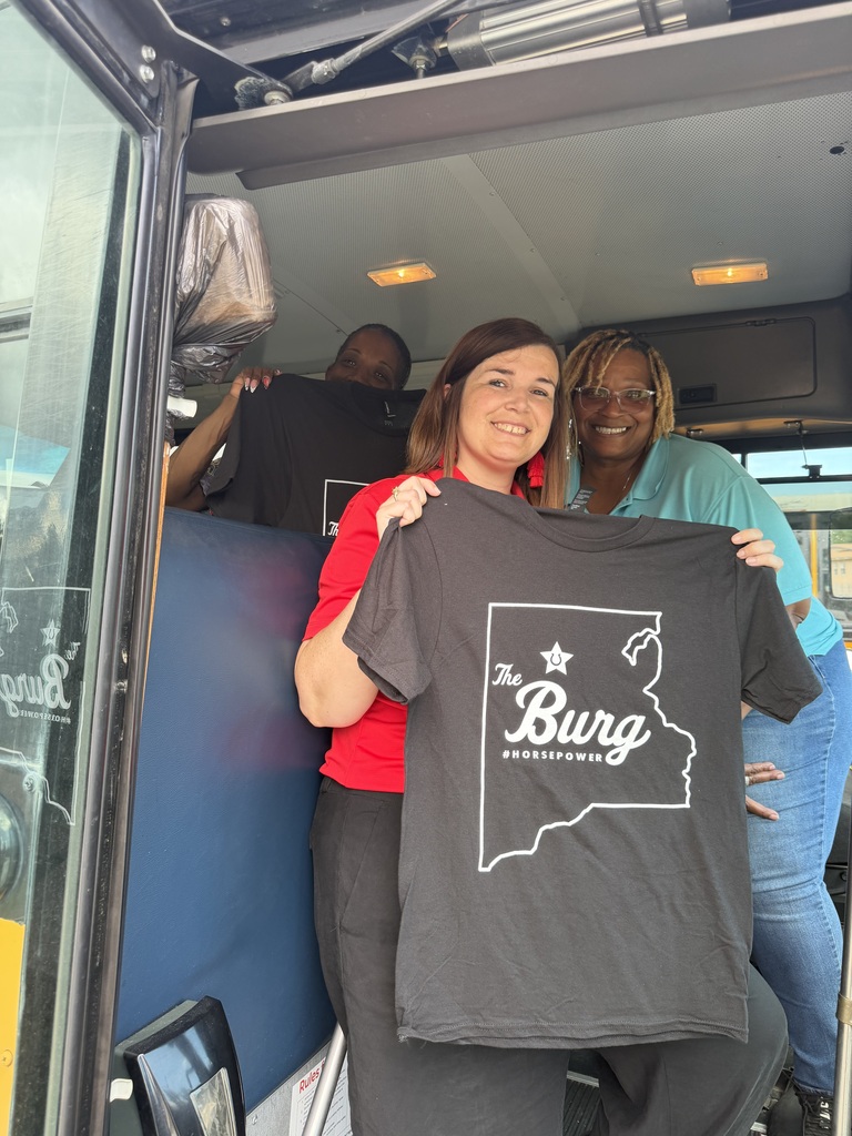 Two women hold a shirt with "The Burg" and a Missouri map outline inside a bus.