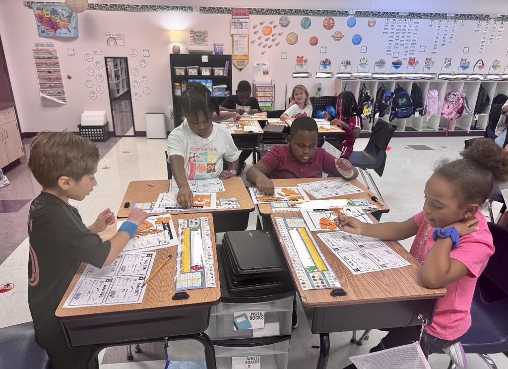 Children in a classroom, seated at desks. One has a bracelet. White walls, colorful decorations, and lockers.