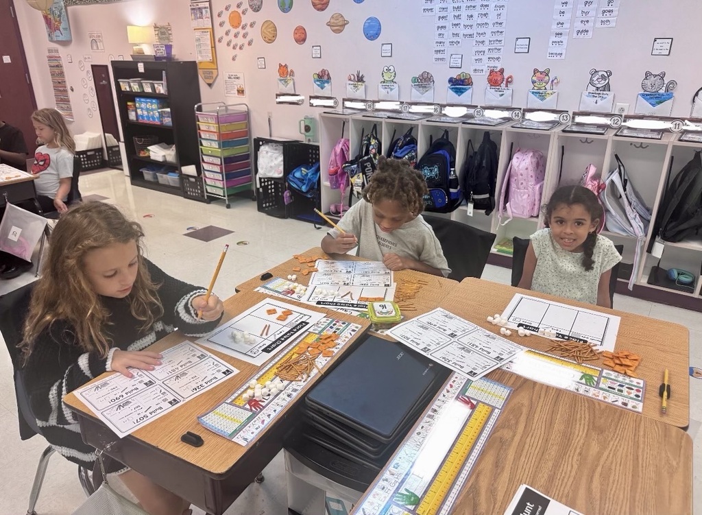 Three young children sit at a table, working on school projects. A few papers are on the table, and a few are in their hands.