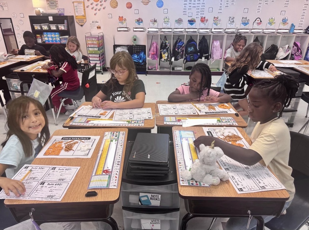 Students seated at tables with papers and toys, smiling. The room has bright decorations and shelves.