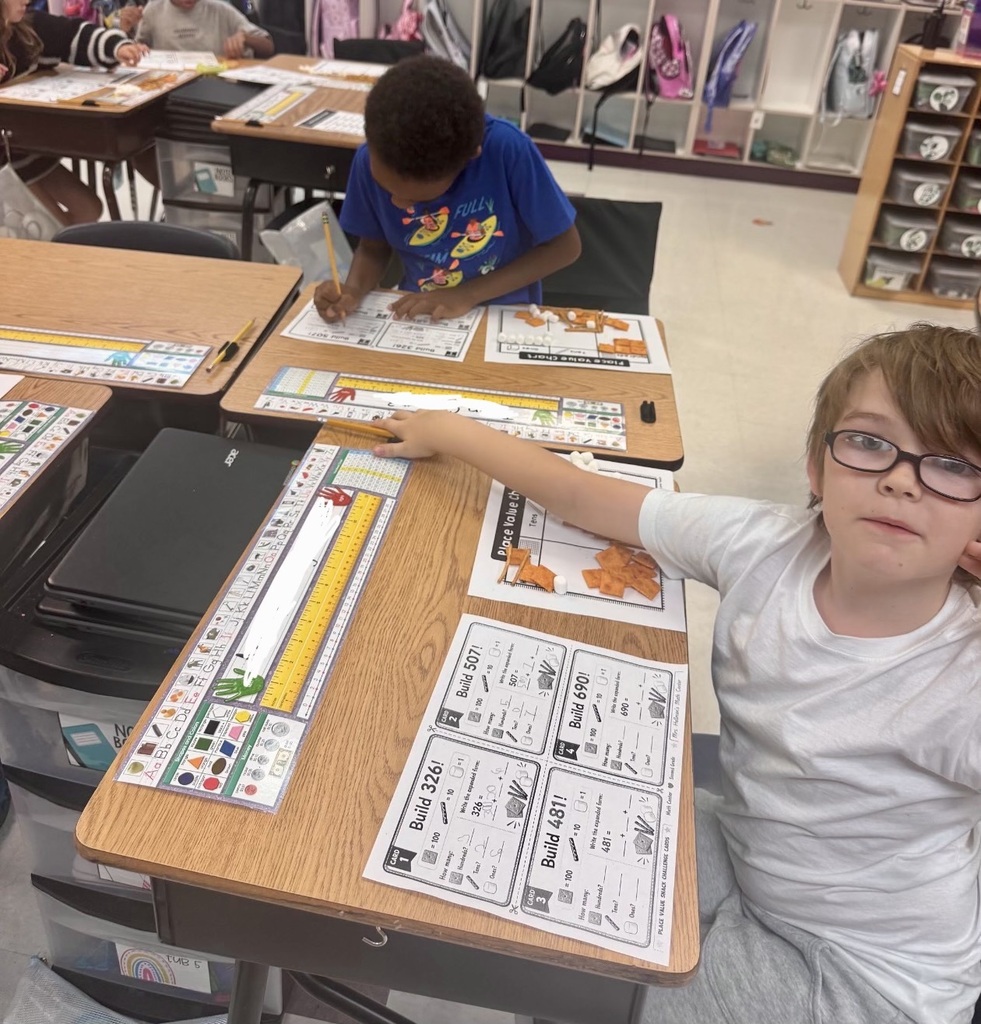 In a classroom, children sit at desks, one wearing glasses and the other using a pencil. Papers and pencils are on the desks.