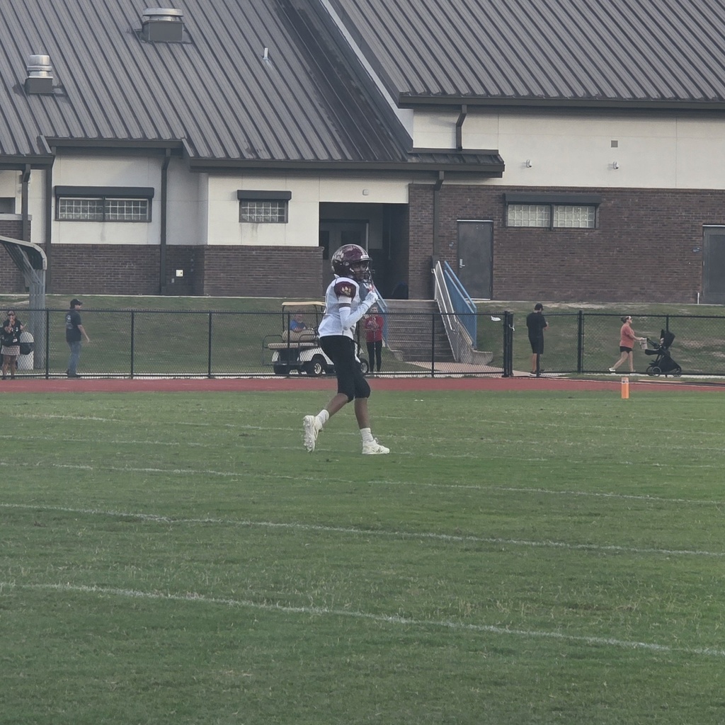 A football player running across a field in front of a building with a black roof.