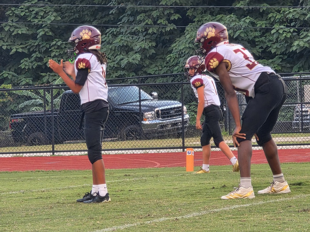 Three football players on a field. One player holds a ball. Behind them, a fence, trees, and a pickup truck.