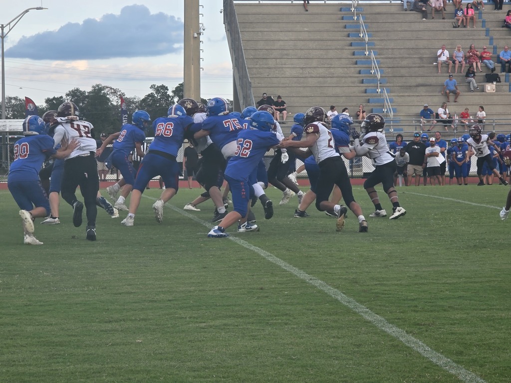 Football players in blue uniforms play on a field with a white line and bleachers filled with spectators.