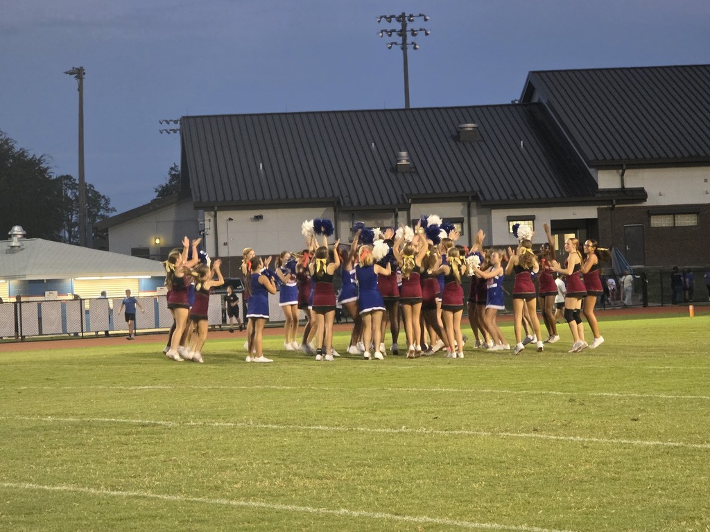 A group of cheerleaders with pom-poms on a football field at dusk, with a building in the background.