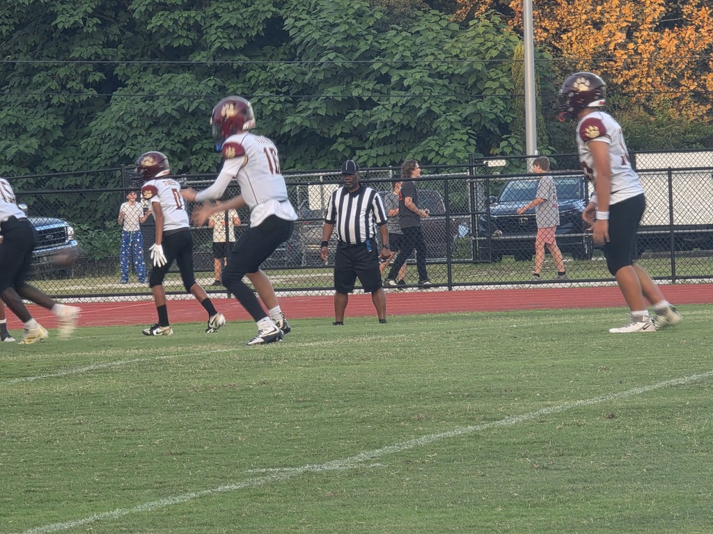 Players on a field, wearing helmets and sneakers. A referee is in the middle. Background includes trees and parked vehicles.