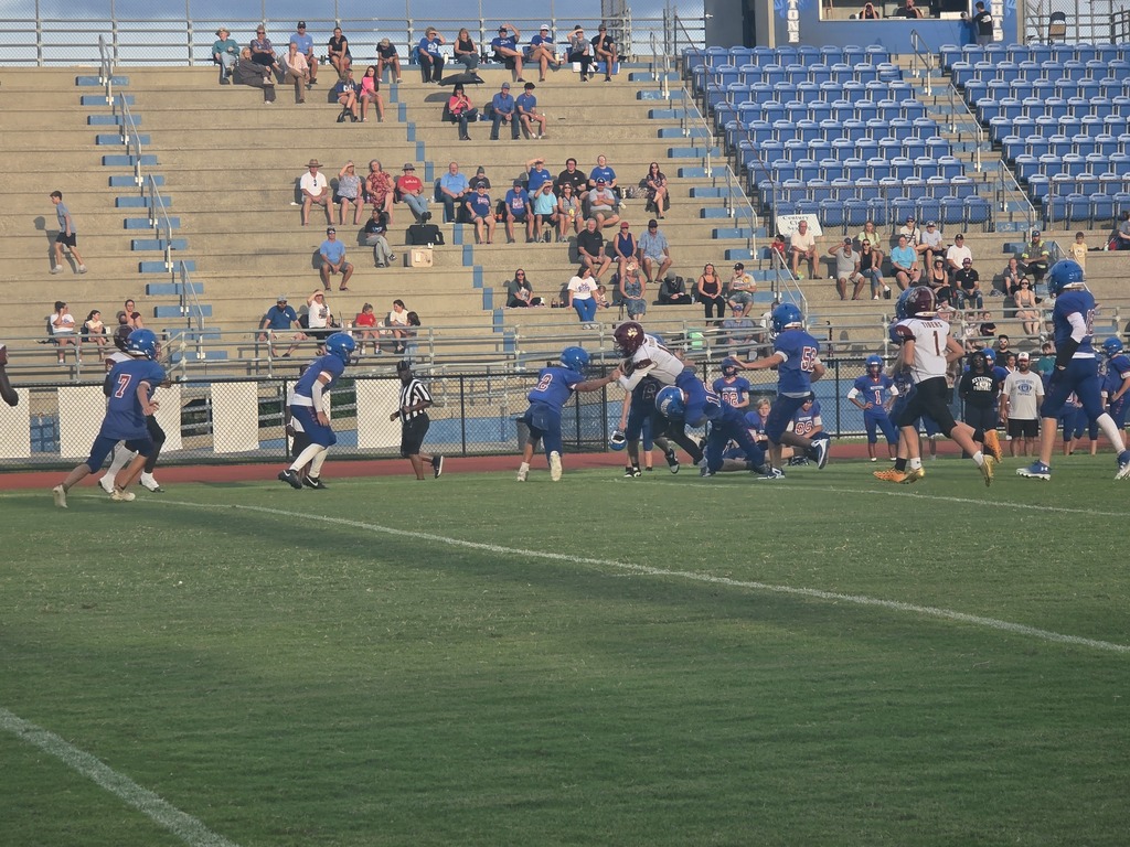 Football game in progress with players in blue uniforms on the field. Spectators seated on blue benches watch the action.